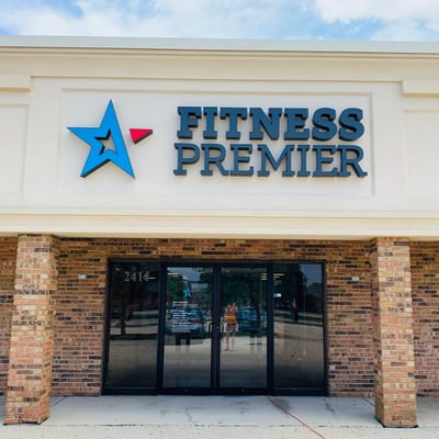 Facade of fitness premier gym with a star logo above clear glass doors, surrounded by a brick wall under a clear sky.