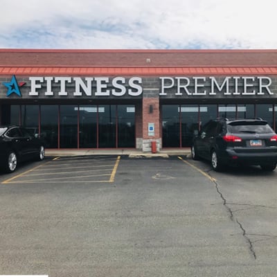 Exterior view of "fitness premier" gym with a clear storefront and cars parked in front.