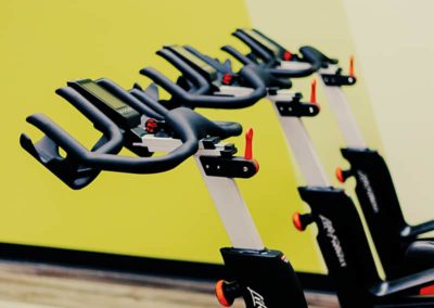 A row of stationary exercise bikes with adjustable handlebars in a gym with a yellow wall.