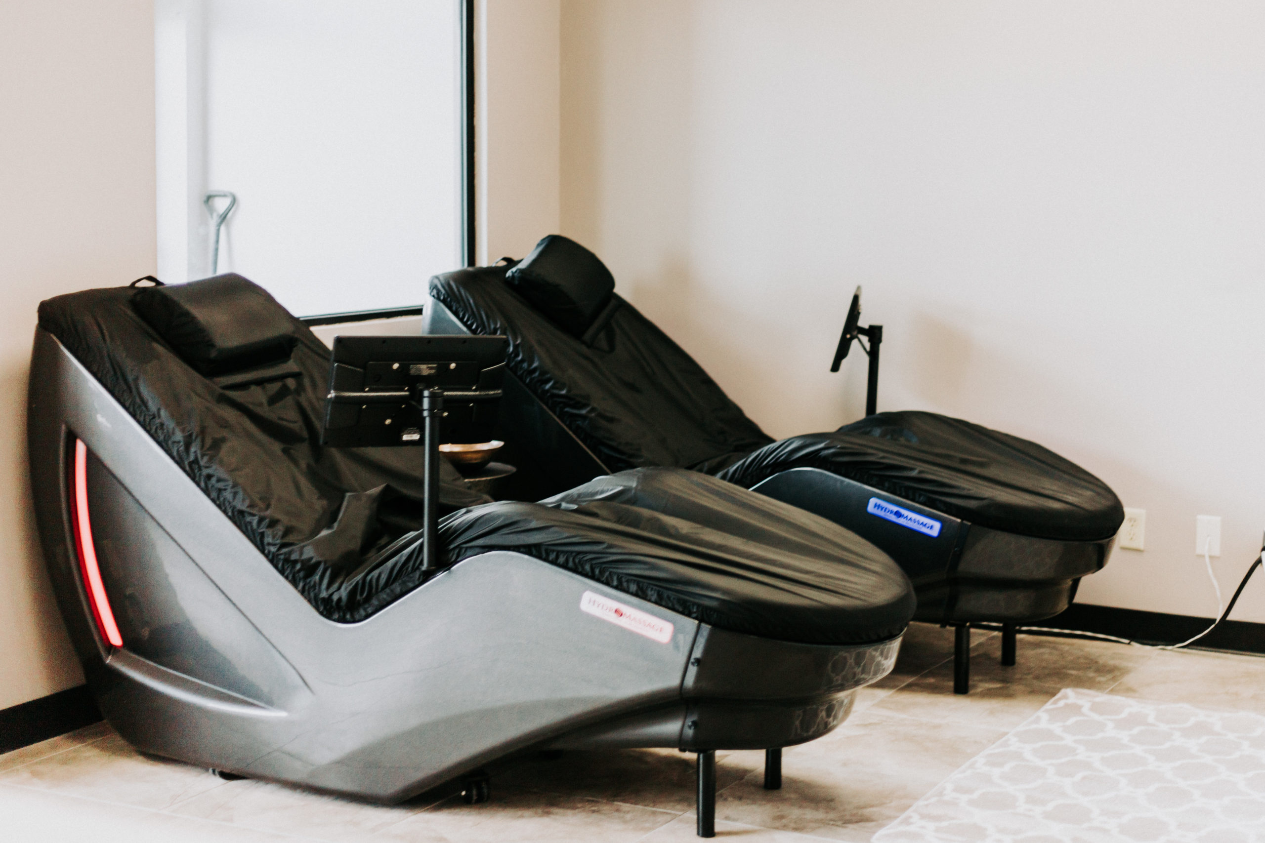 Two black massage chairs in a modern room with gray walls and a white, patterned rug, featuring blue screen interfaces.