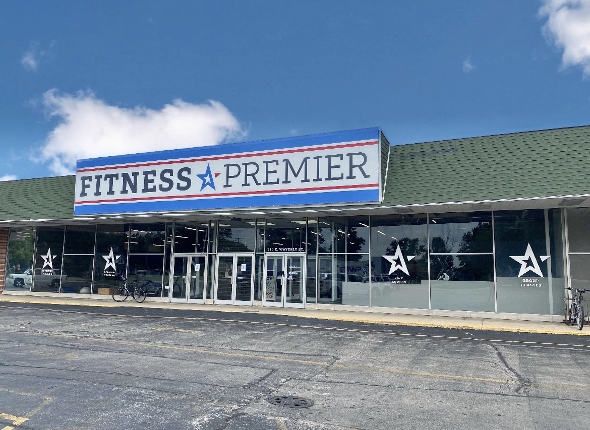 Exterior of fitness premier gym with blue and red signage above the entrance, parked bicycles outside under a clear sky.