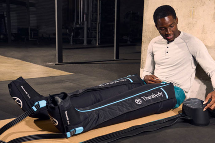 Man in gym clothes sitting on the floor, using a portable massage device next to a large black and blue gym bag.