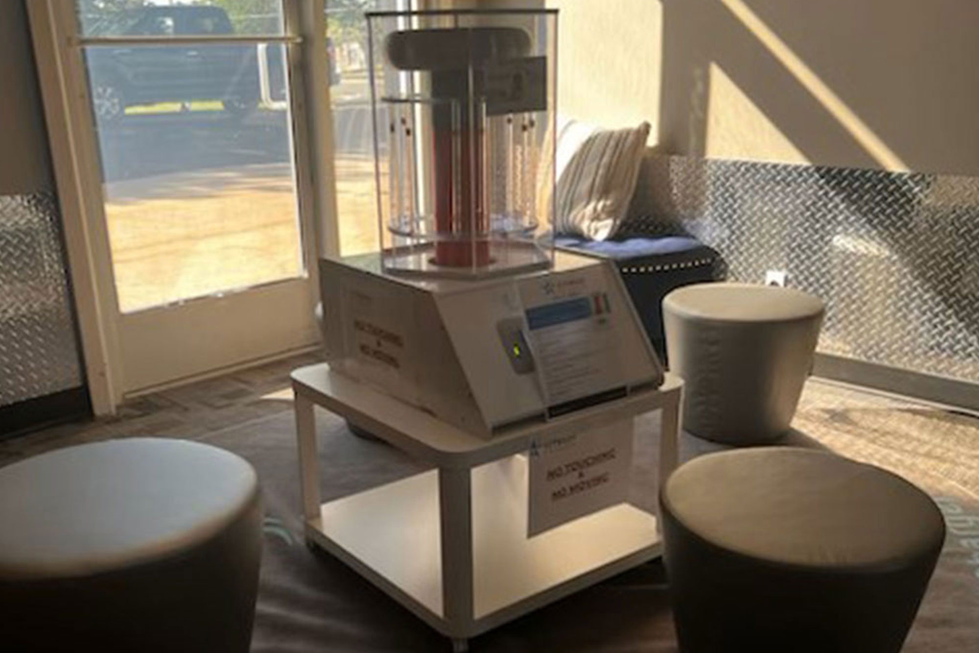 A medium-sized disinfecting machine, encased in a clear box, sits on a white table in a well-lit room with a window, seating, and metallic wall detailing.