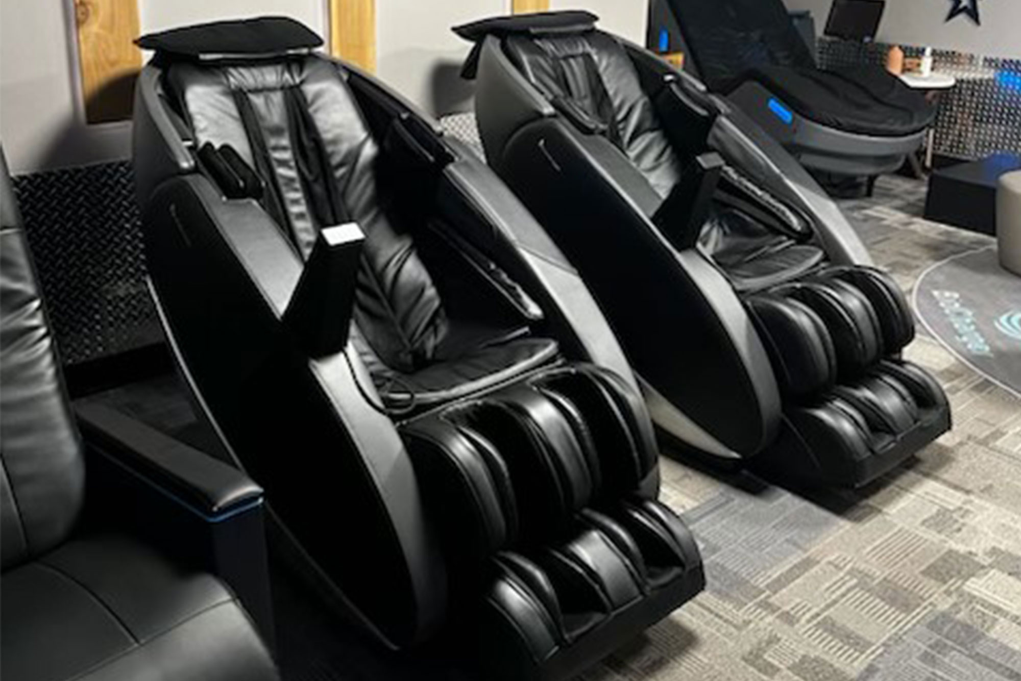 Two black massage chairs are positioned side by side in a modern room with carpeted flooring and a sleek design. The chairs feature multiple massage rollers and footrests.