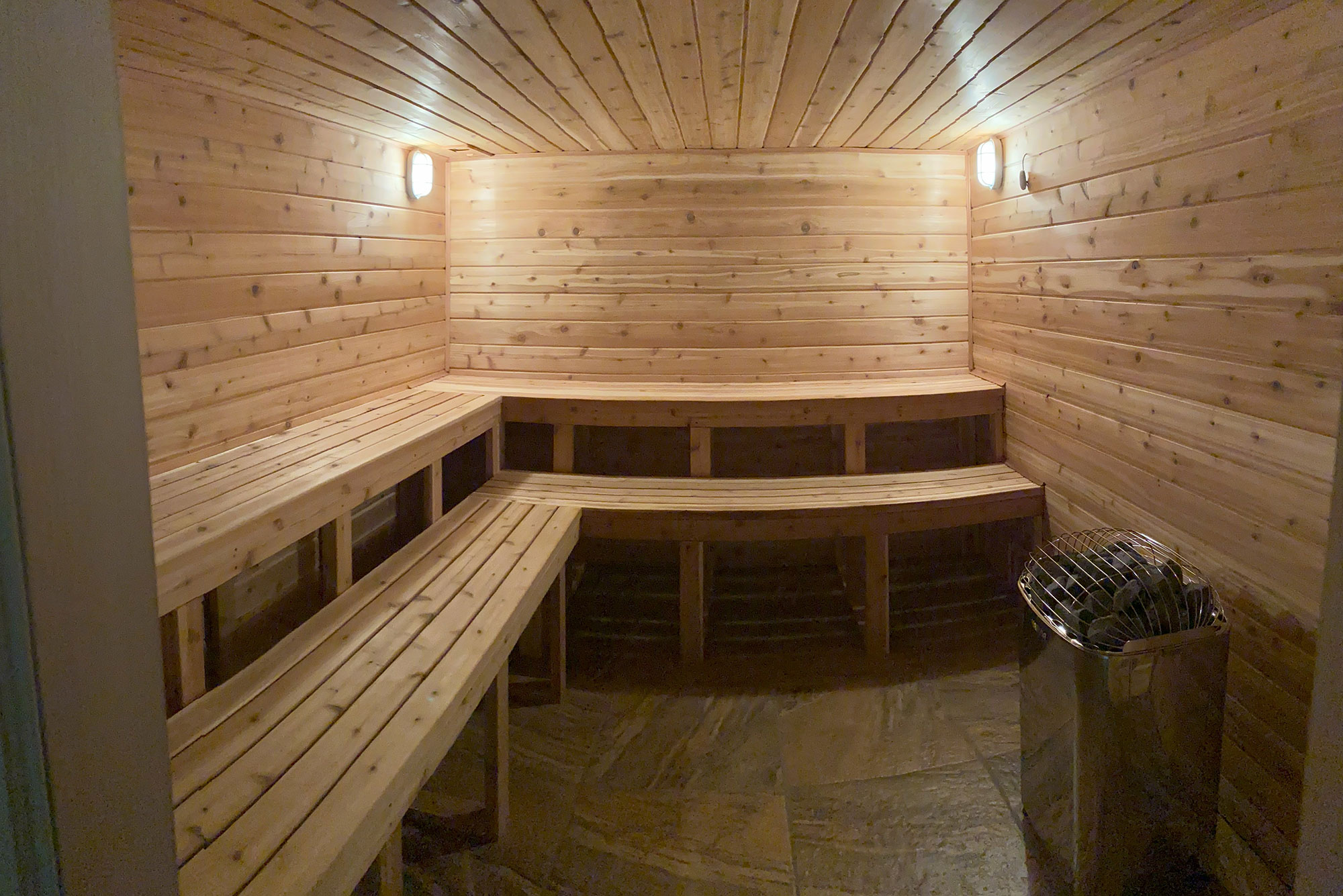 An empty, wooden sauna room with benches along the walls, lit by two wall-mounted lights, and a metal heater in the corner.