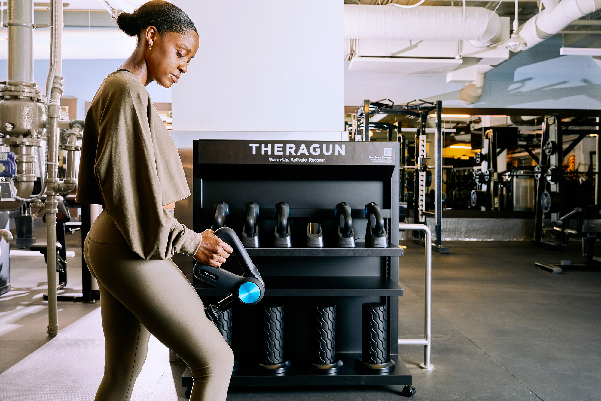 A woman in athletic wear uses a Theragun in a gym, standing near a rack of other Theragun devices against a backdrop of gym equipment and fitness machines.