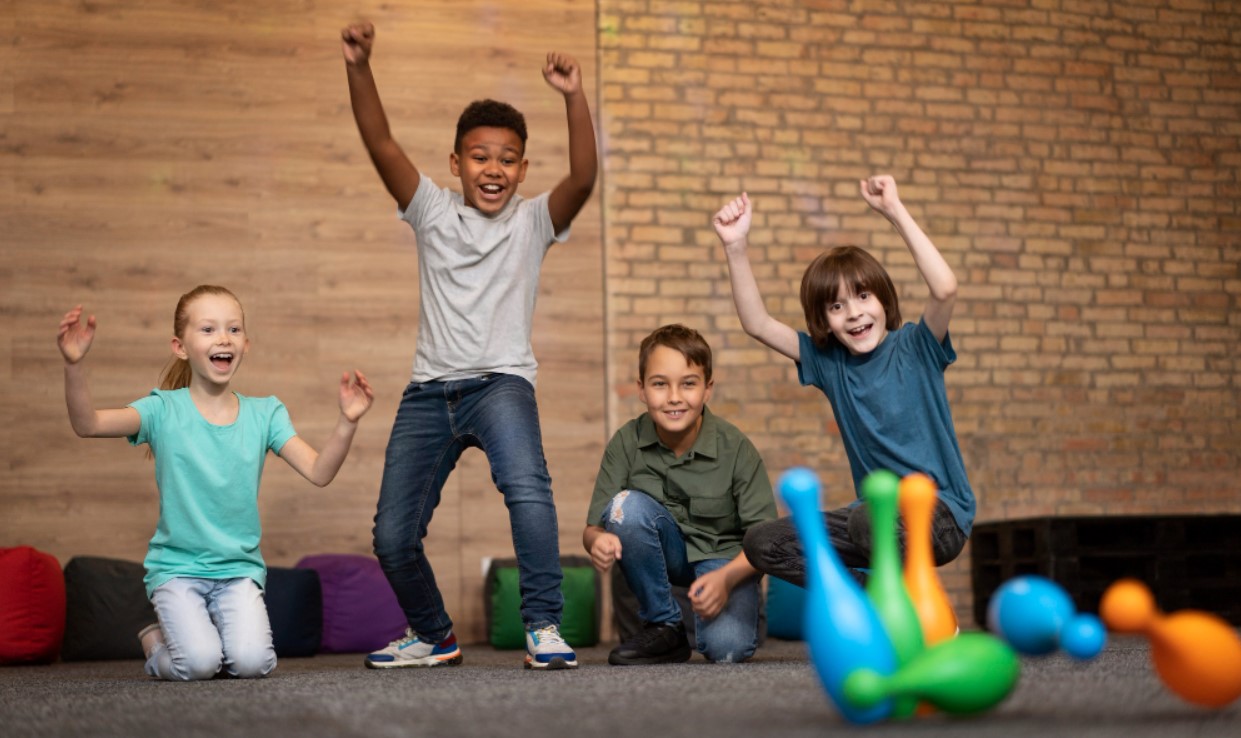 Four children cheer and raise their arms while playing with colorful bowling pins indoors.