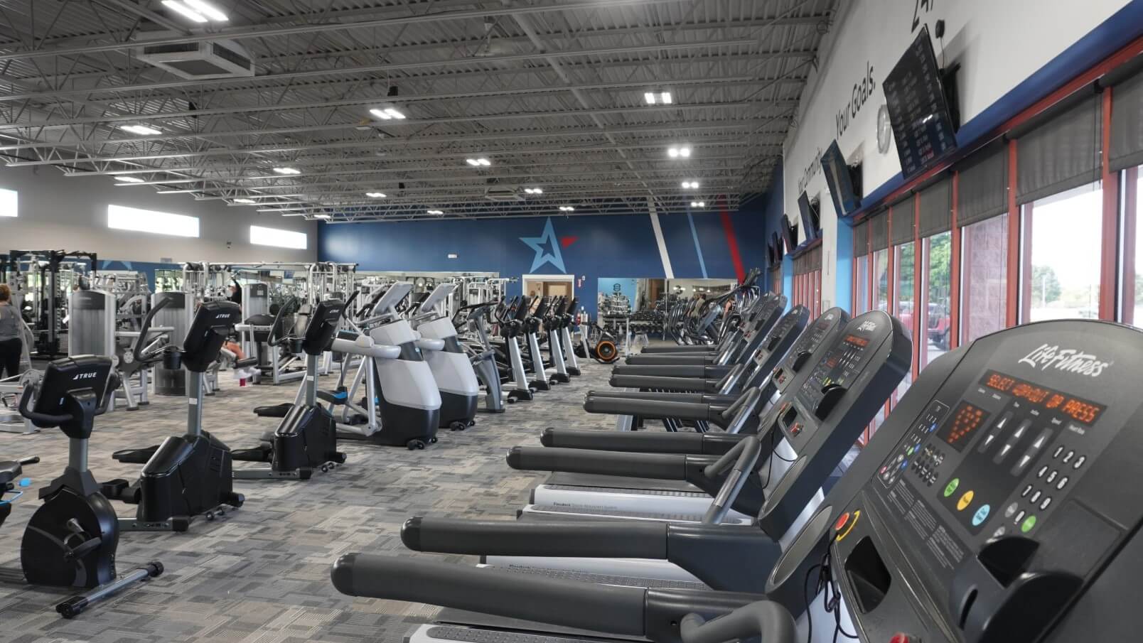 A spacious gym with treadmills, exercise bikes, and weight machines lined up in rows. Large windows allow natural light to fill the room.