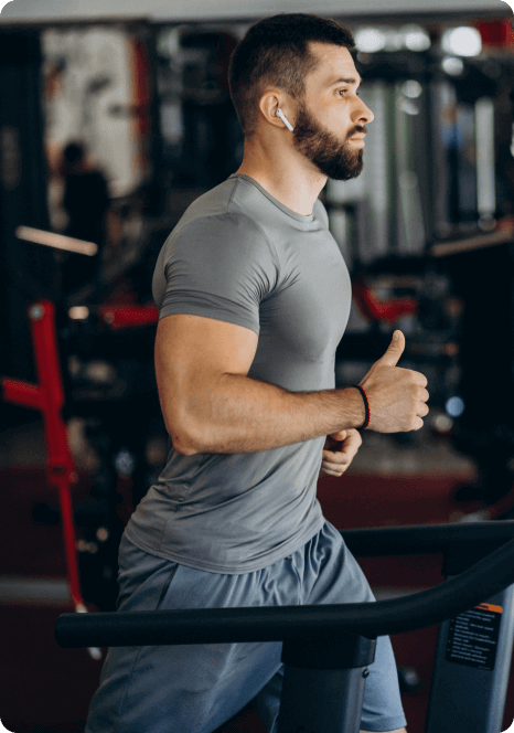 Man with a beard running on a treadmill in a gym, wearing a gray shirt and earphones.